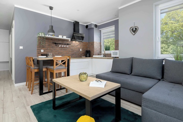 A modern living room connected to a kitchenette, featuring a gray corner sofa and a wooden coffee table on a dark rug. In the background, you can see a large window that lets in natural light.