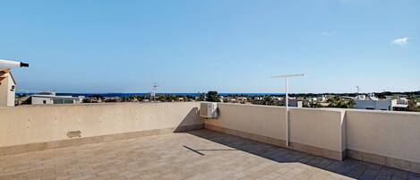A rooftop terrace with a clear view of the sea, a concrete floor, and a few buildings in the background.