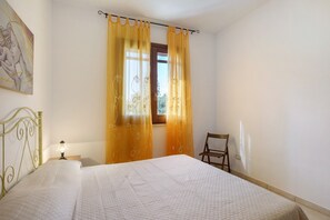 A bedroom with a bed, curtains, a chair, a nightstand, and a wall-mounted lamp. - Apartment 'Giallo' with Terrace, Wi-Fi and Air Conditioning (San Vito Lo Capo)