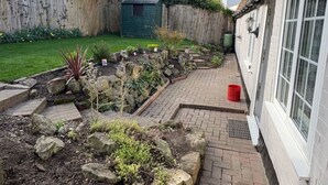 Un jardín con un sendero de piedra, un cobertizo pequeño y una casa de fachada blanca y ventanas amplias.
