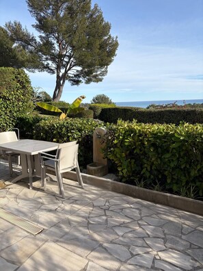 Um pátio externo com mesa e cadeiras, rodeado por vegetação e vista para o mar.