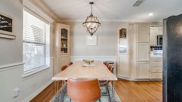 A dining room with a wooden table, chairs, and a chandelier. There are cabinets and a refrigerator in the background.