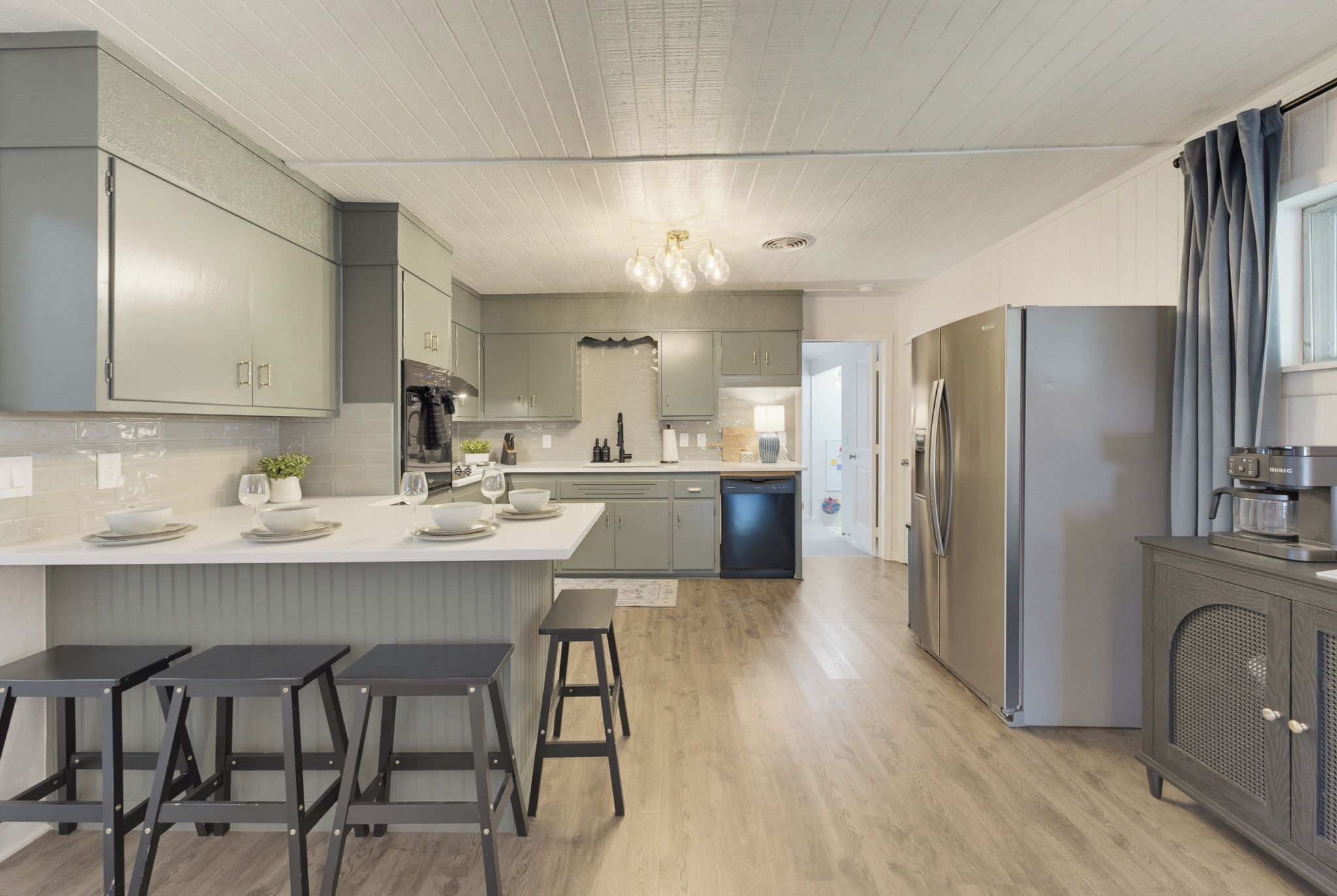 A modern kitchen with grey cabinets, a central island with bar stools, a built-in dishwasher, and a stainless steel refrigerator.