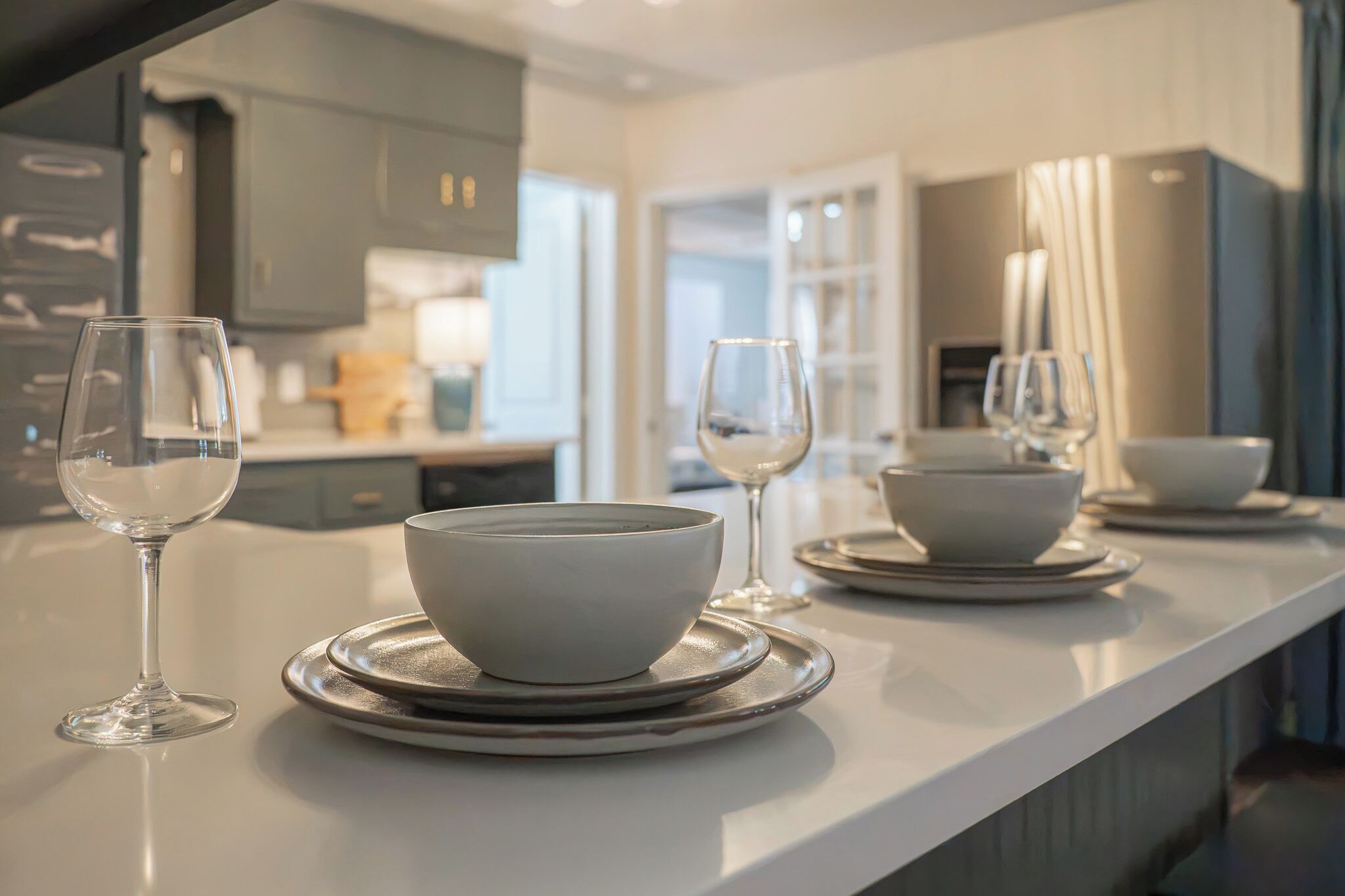 A modern kitchen with a white countertop, two bowls on plates, and two wine glasses.