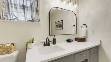 Une salle de bain avec un lavabo blanc, un miroir avec éclairage intégré, un porte-serviettes et une petite plante.