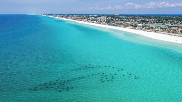 Eine Luftaufnahme einer Küstenregion mit klarem türkisblauem Meer, einem weißen Sandstrand und einer dicht bebauten Stadt im Hintergrund.