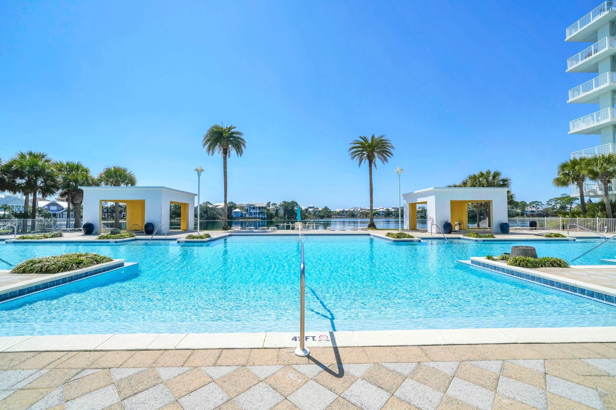 Piscina azul cristalina com plataforma de mergulho, palmeiras e um edifício com varandas ao fundo.