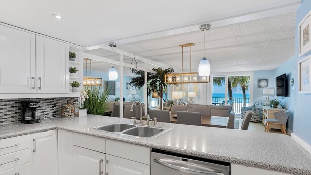 A modern kitchen with a dishwasher, double sink, and a dining area with a view of the ocean. - Coastal Luxury at Sand Castle II 205 (Indian Shores)