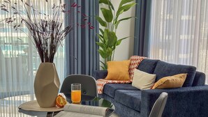 Een moderne woonkamer met een blauwe bank, een glazen tafel met een boek en een glas sinaasappelsap, en een vaas met gedroogde planten.
