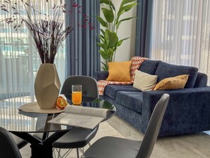 Ein modernes Wohnzimmer mit einem blauen Sofa, einem Glastisch mit einem Buch und einem Glas Orangensaft sowie einer Vase mit Trockenpflanzen.