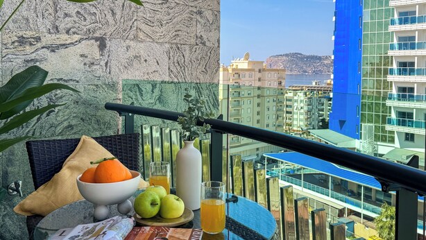 Een balkon met een tafel, daarop fruit, een sapglas, een tijdschrift en een bloemenvaas.