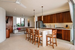 A modern kitchen with wooden cabinets, a central island with chairs, and a dining area with a sofa and window. - Luxury Livving Steps from La Ropa - 2Br 2 Bath (Zihuatanejo)