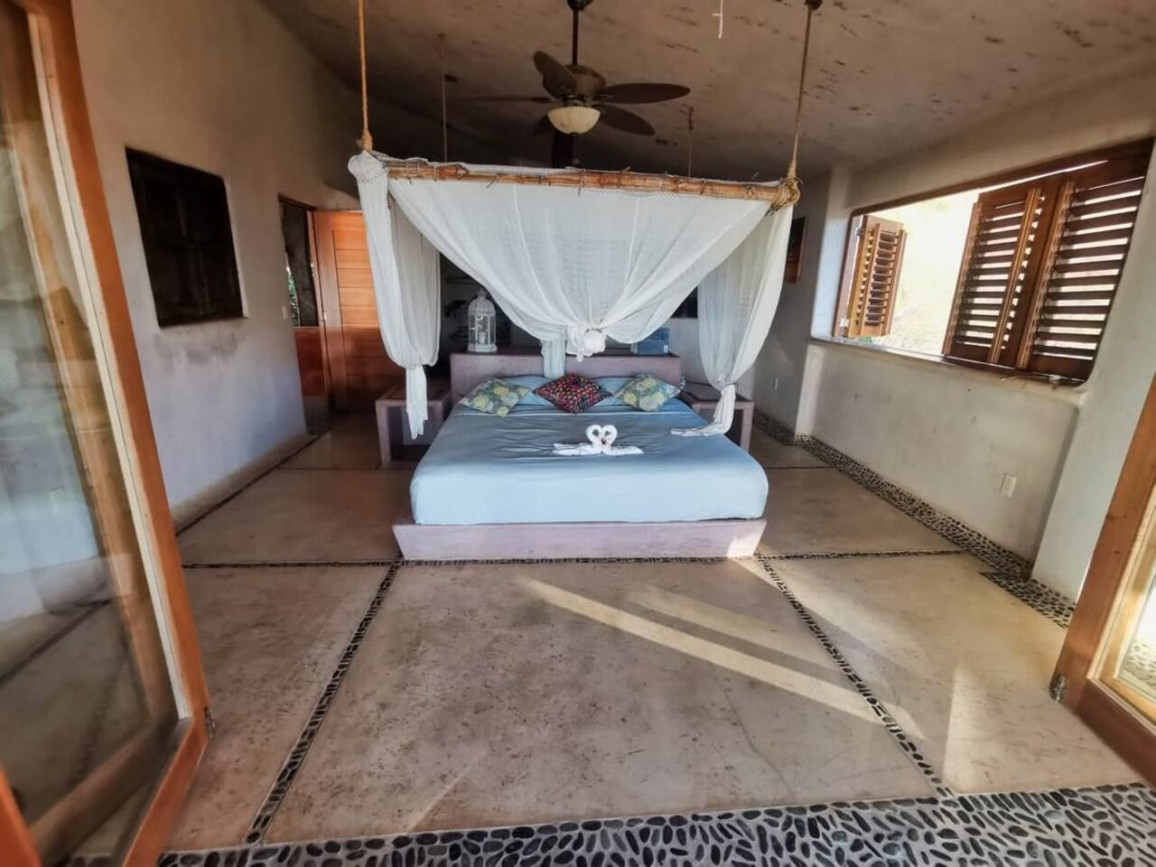 Une chambre avec un lit sous un baldaquin, un ventilateur de plafond et une fenêtre avec des volets.