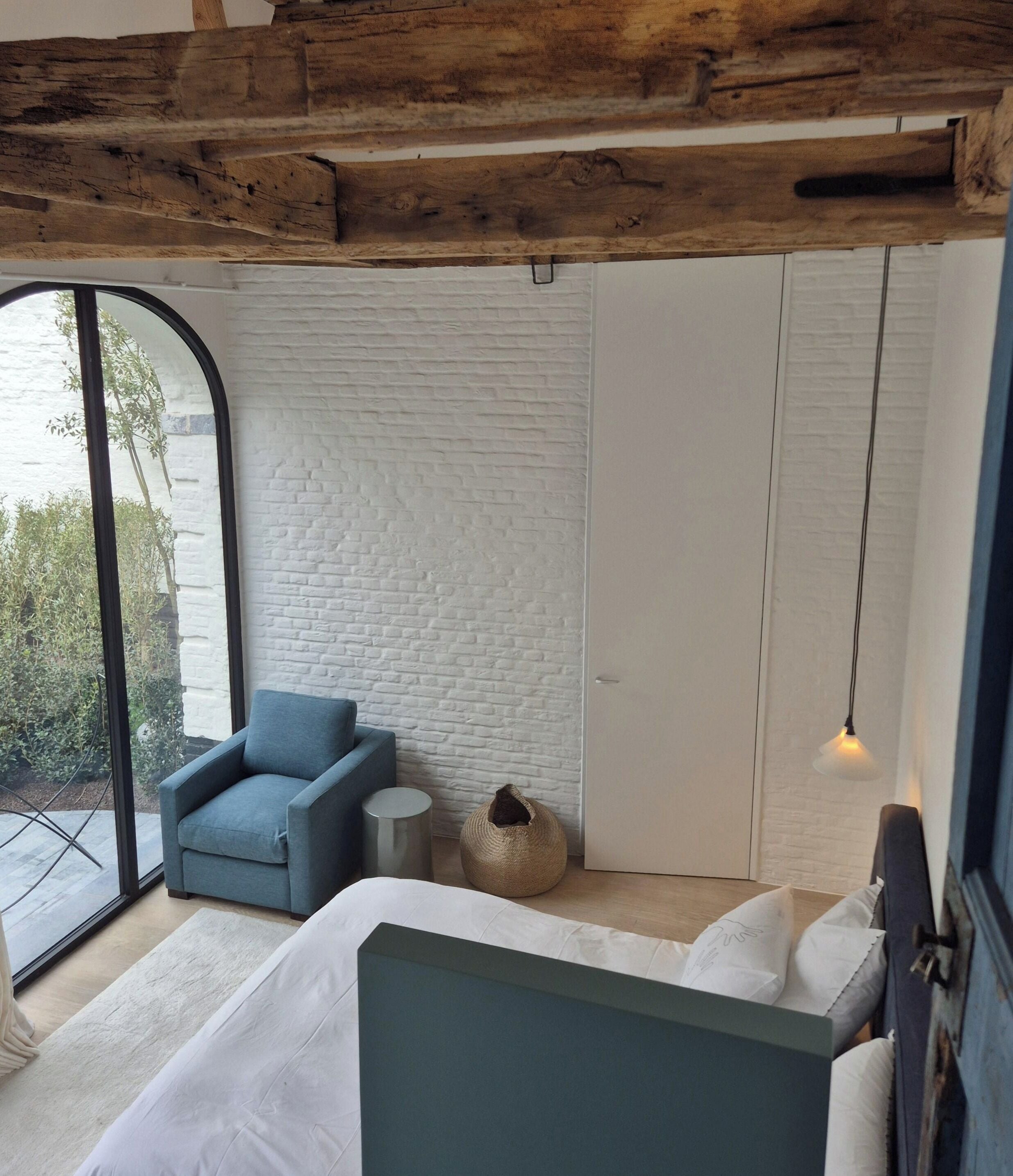 A bedroom with a bed, a blue armchair, a round woven stool, and a large window.