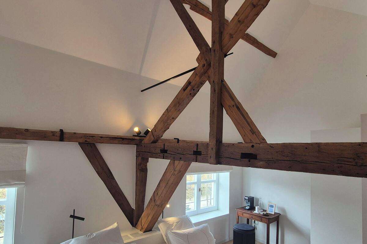 A bedroom with exposed wooden beams, a bed with white bedding, a small table with a lamp, and a window with daylight.