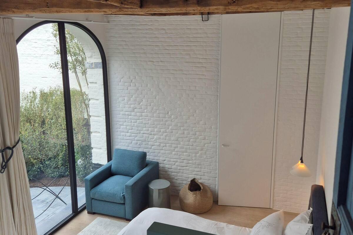 A bedroom with a bed, a chair, a wardrobe, a round wicker basket, and a view through a large window.
