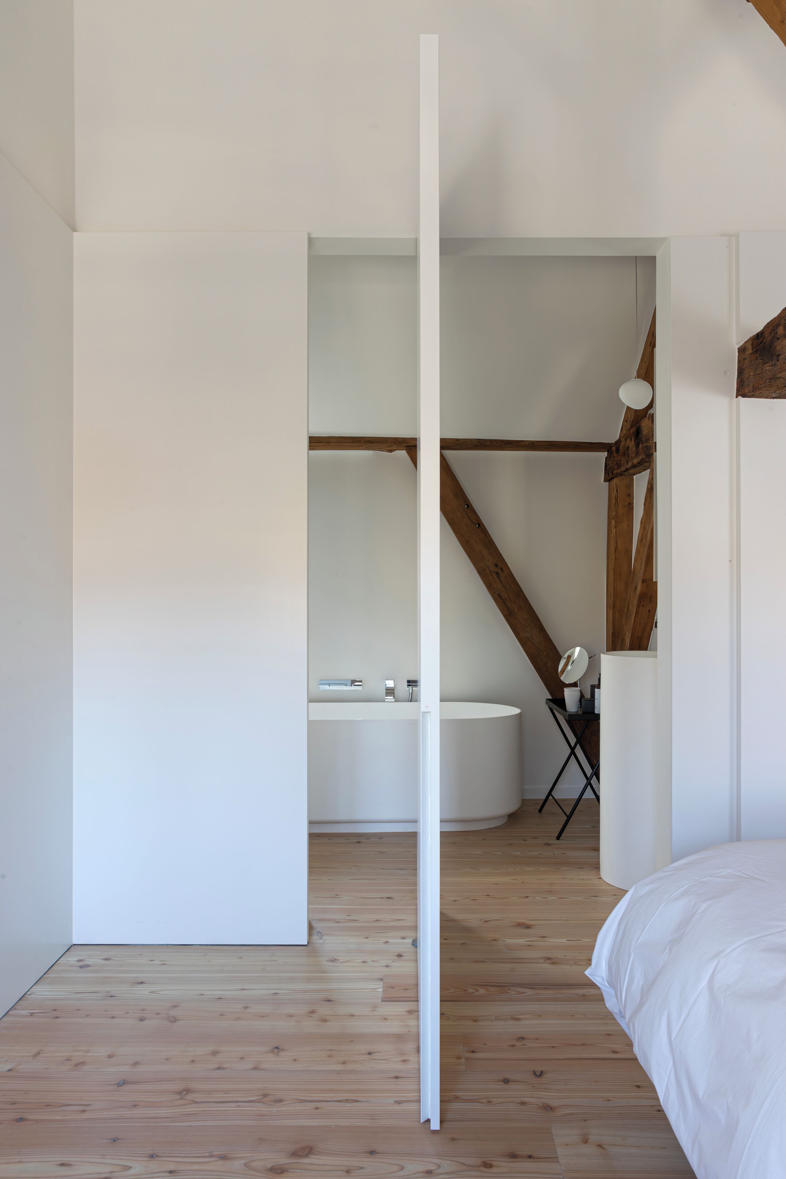 A bedroom with a wooden floor, a white bed, a white wall, and a wooden ceiling beam.