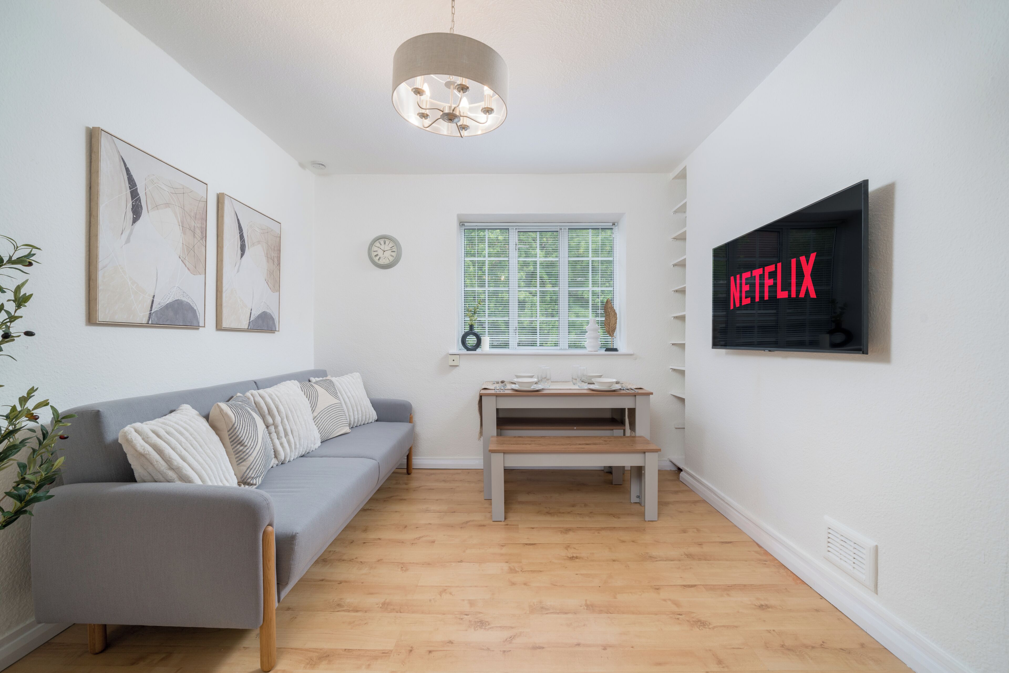 A modern living room with a grey sofa, a wooden coffee table, a window with blinds, a wall-mounted TV, and abstract wall art.