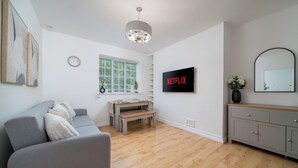 Een moderne woonkamer met een grijze bank, houten vloeren, een aan de muur bevestigde televisie en een raam met zonwering.