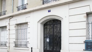 A white building with a black ornate door, a small balcony with metal railings, and a number '7' sign above the door. - Bright studio apartment on the outskirts of Paris (Clichy)