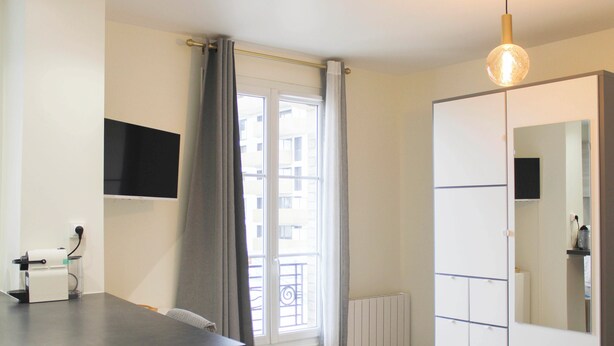A modern room with a flat-screen TV, a radiator, a window with curtains, a wardrobe, and a pendant light. - Bright studio apartment on the outskirts of Paris (Clichy)
