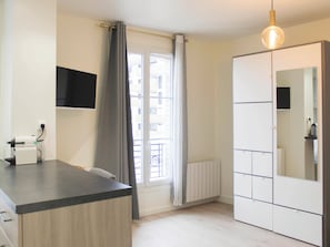 Quarto moderno com televisão de tela plana, radiador, janela com cortinas, guarda-roupa e luminária pendente.