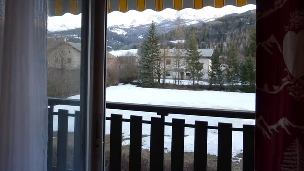 Et værelse med udsigt til et sneklædt landskab, en balkon med gelænder af træ og et vindue med gule gardiner.
