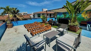 Un espace piscine aménagé sur le toit, avec des chaises longues et des tables, entouré de plantes en pot et surplombant un ciel d’un bleu limpide.