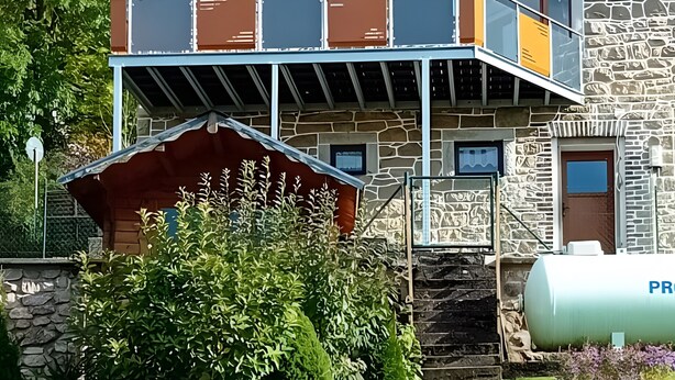Un edificio a due piani con tetto in tegole rosse, facciata in pietra e un balcone con ringhiere in metallo.