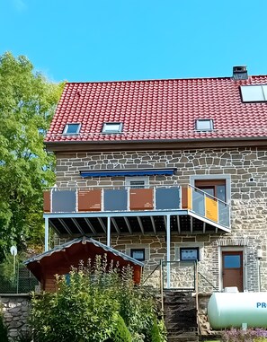 Un bâtiment à deux étages, doté d’un toit recouvert de tuiles rouges, d’une façade en pierre et d’un balcon orné de rampes en métal.