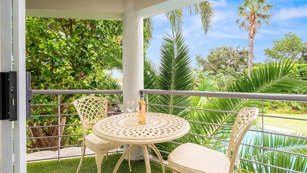 A covered outdoor area with a round table and chairs, a bottle of wine, and a view of a pool and palm trees. - The Oceana (Cape Town)