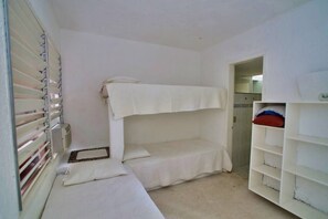 Ein kleines Zimmer mit einem Einzelbett und einem Etagenbett, einem Heizkörper, einem Regal mit Kissen und einer Tür zu einem anderen Raum.