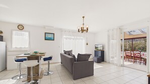 En moderne stue med en grå sofa, spiseplads med bord og stole, et køkkenområde og et stort vindue med gardiner.