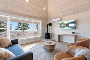 A cozy living room with a sofa, coffee table, and leather chairs, a fireplace, and a large window with a view of trees.