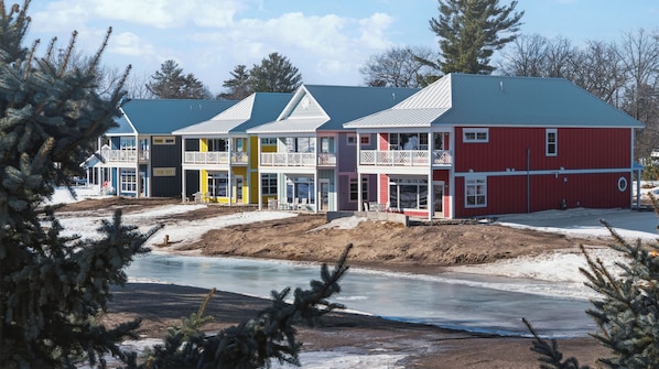 A row of multi-story buildings with balconies, a frozen river in the foreground, and trees in the background. - Mille Lacs Suite | King Bed & Soaking Tub (Garrison)