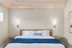 A bedroom with a wooden ceiling, a bed with white bedding and a blue blanket, two bedside lamps, and a round side table with a patterned cushion. - Modern Mille Lacs Suite | Sleeps 6 (Garrison)