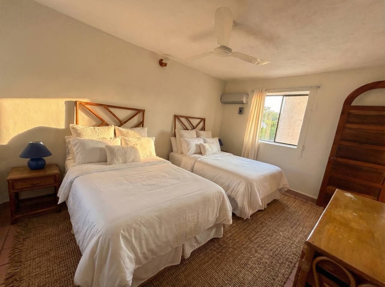 A bedroom with two beds, a ceiling fan, a window with curtains, and a wooden table.