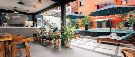 A reception area with a reception desk, chairs, and a counter. There is a pool area with lounge chairs and umbrellas.
