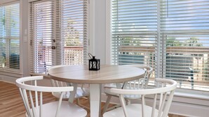 A room with a round table and four white chairs, large windows with blinds, and a view of greenery outside. - Sand Casas (Port St. Joe)