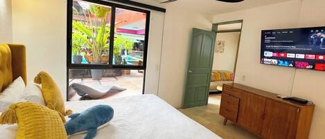 A bedroom with a bed, a wooden dresser, a television, and a sliding glass door leading to an outdoor area with plants.