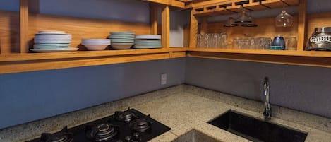 Una cocina con estufa a gas, un lavamanos, repisas de madera con vajilla y un soporte para copas de vino.