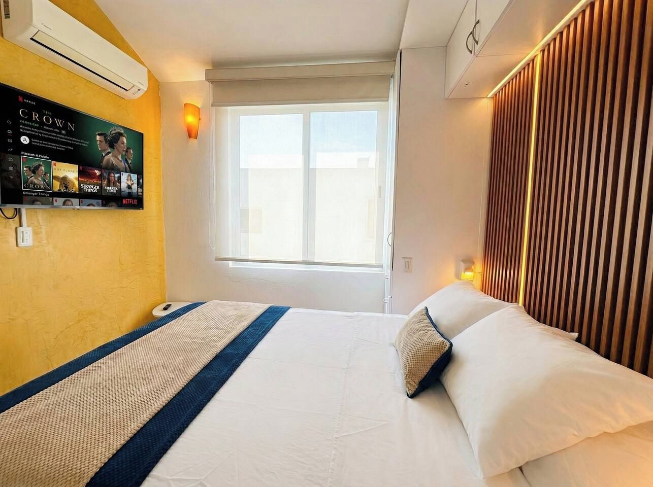 Een hotelkamer met een bed, een aan de muur bevestigde televisie waarop een film draait, een raam met zonwering en een wand met verticale lamellen.