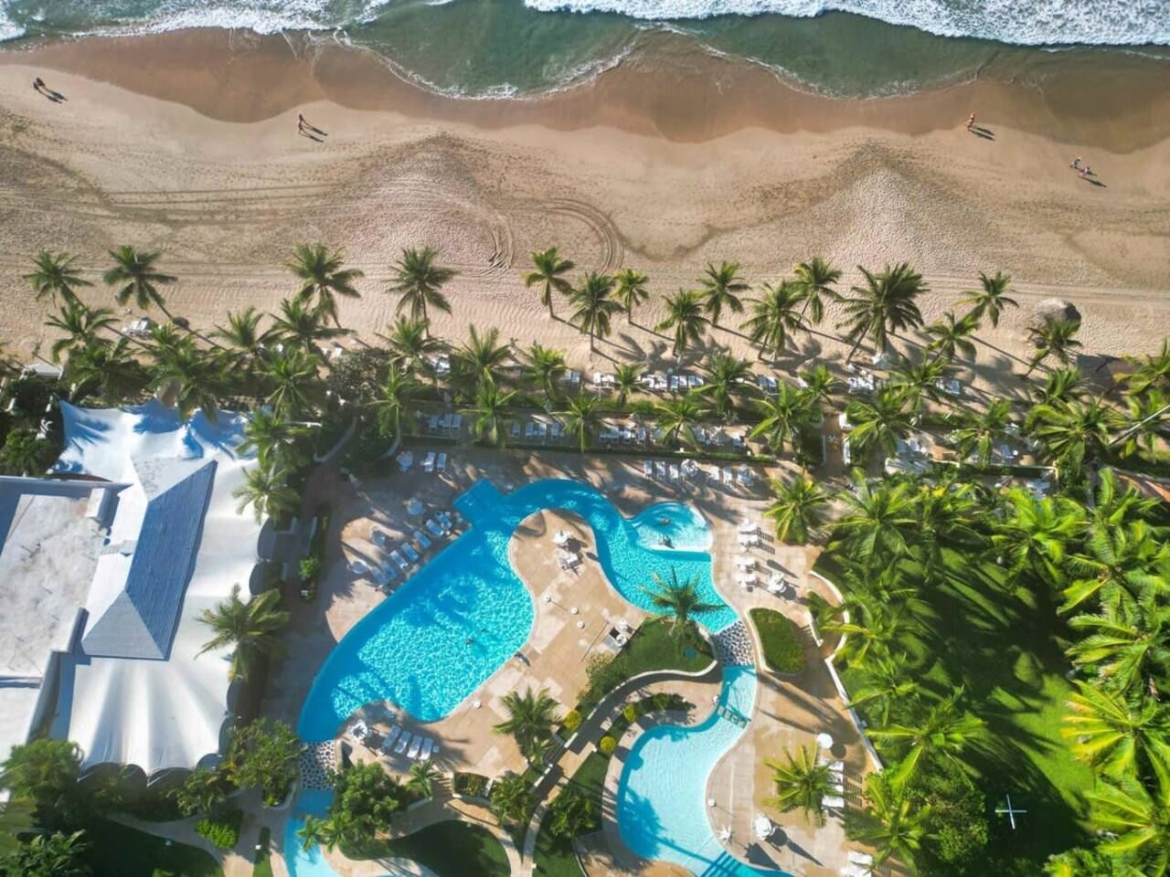 An aerial view of a resort with a large swimming pool, palm trees, and a beach.