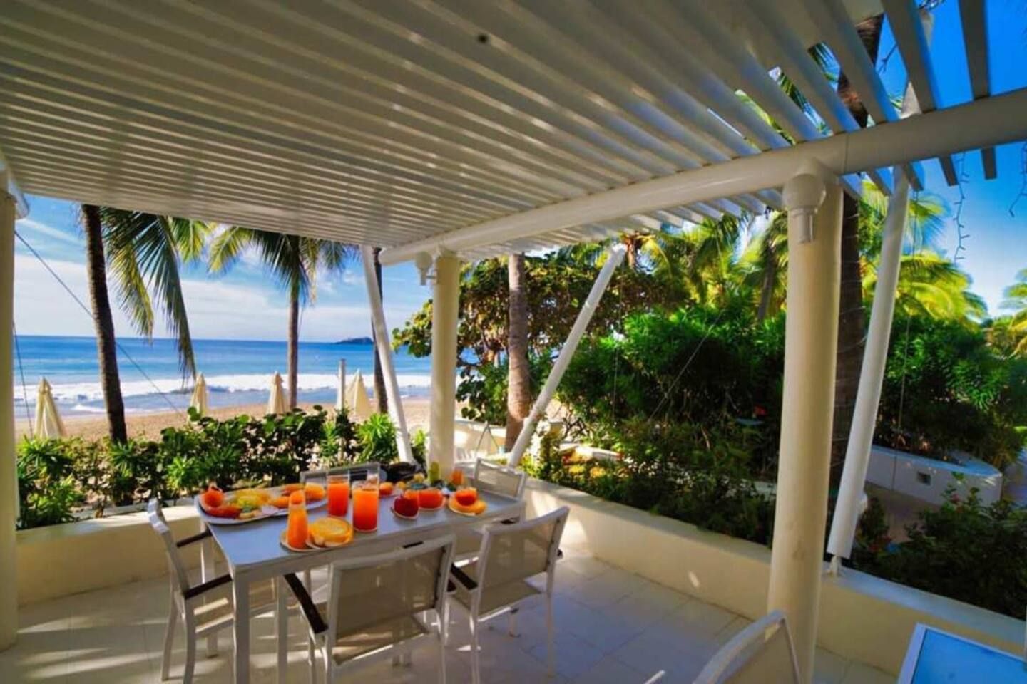 A covered outdoor dining area with a table set for breakfast, overlooking a beach and ocean.