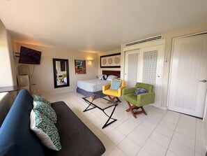 A room with a bed, a sofa, a glass table, two chairs, a TV, and a door with a louvered design. - Elegant Beachfront Studio in Monarca Ixtapa (Ixtapa)