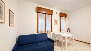 Une pièce avec un canapé bleu, une table à manger blanche avec des chaises, et deux fenêtres avec des rideaux.