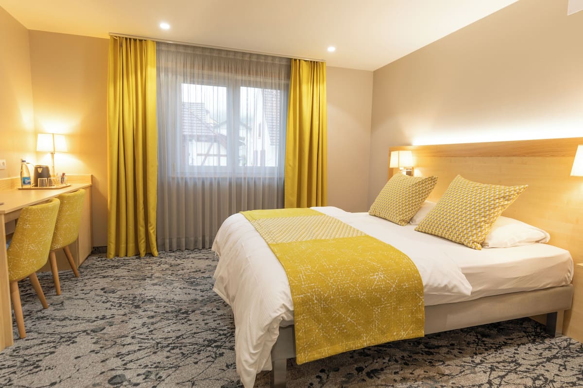 Ein Hotelzimmer mit einem Bett, Nachttischlampen, einem Schreibtisch mit Stühlen und einem Fenster mit Vorhängen.