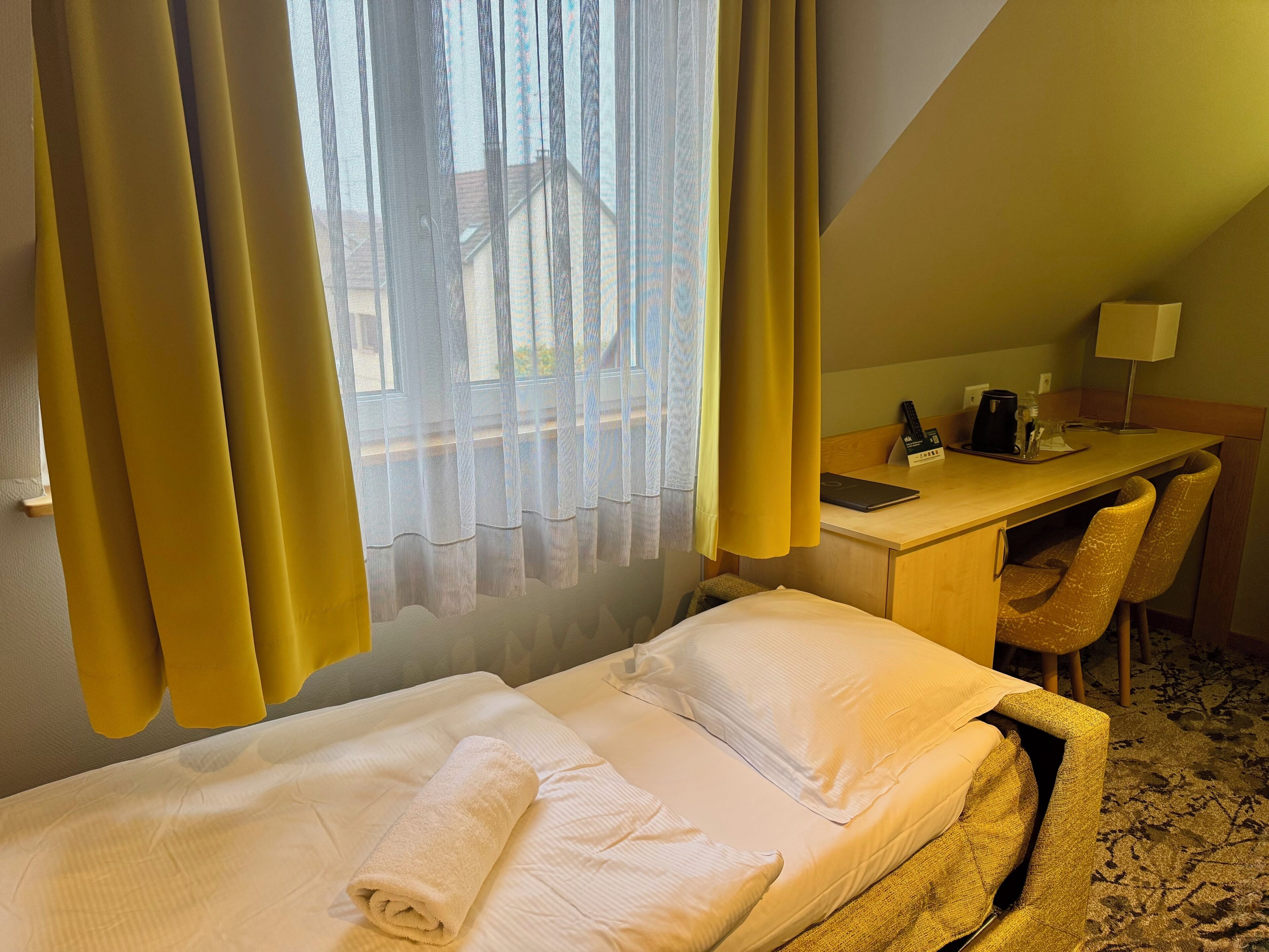 A hotel room with a bed, a desk, a chair, a lamp, and a window with curtains.