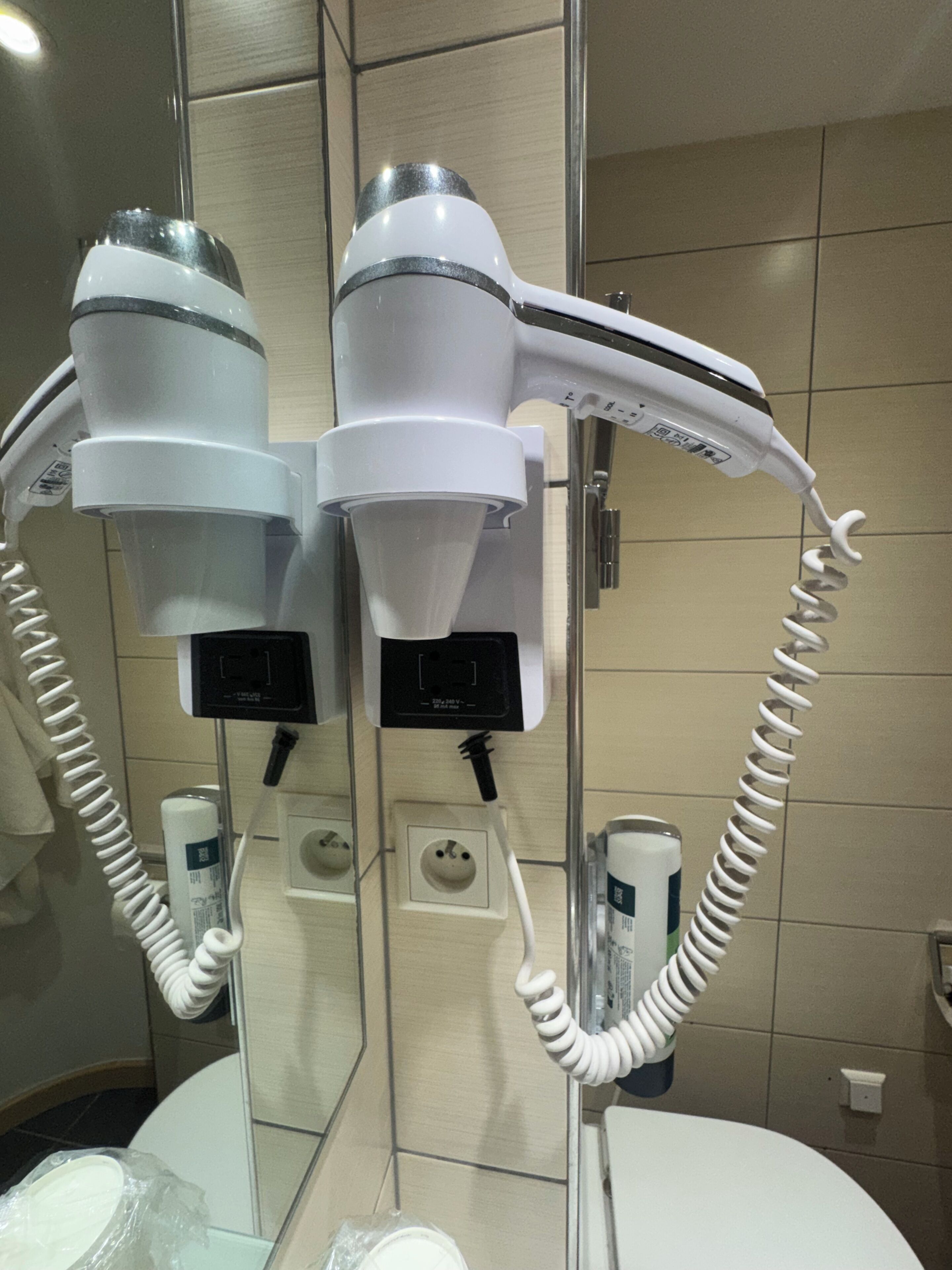 A bathroom with two hair dryers mounted on the wall, a mirror, and a sink with a towel dispenser.