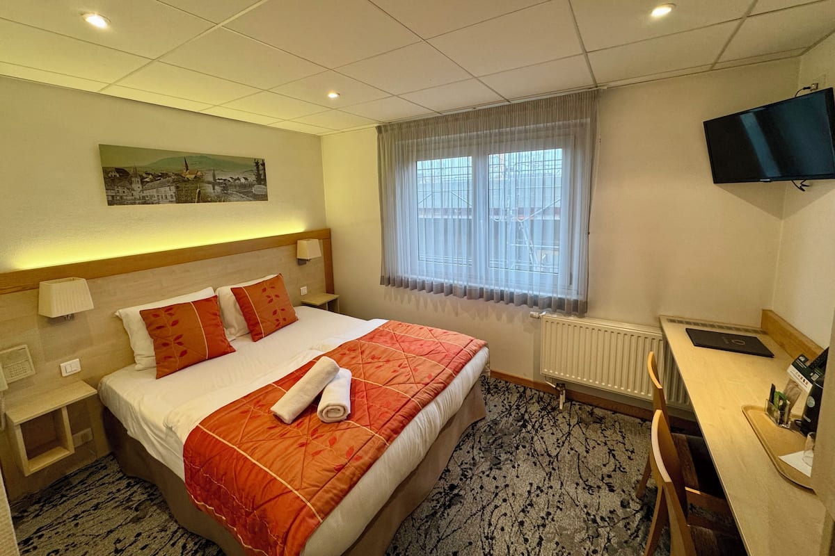Ein Hotelzimmer mit Bett, Schreibtisch, Fernseher, einem Fenster mit Vorhängen und einem Bild an der Wand.